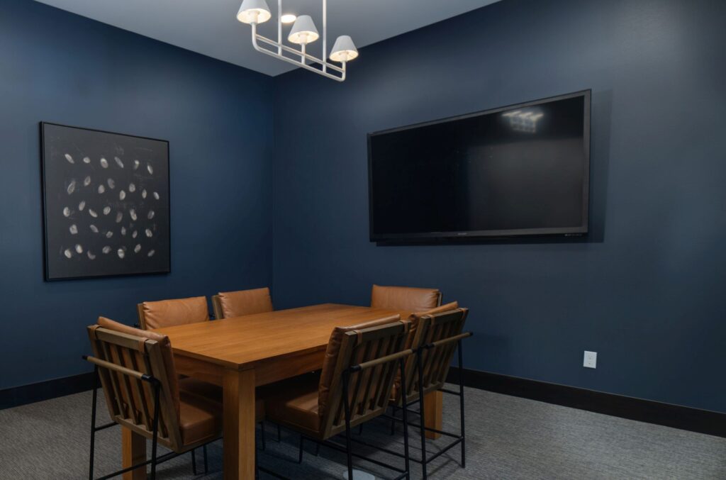 Modern conference room with wood table, leather chairs, whiteboard, and abstract art on a dark blue wall with a mounted television. Office meeting space.