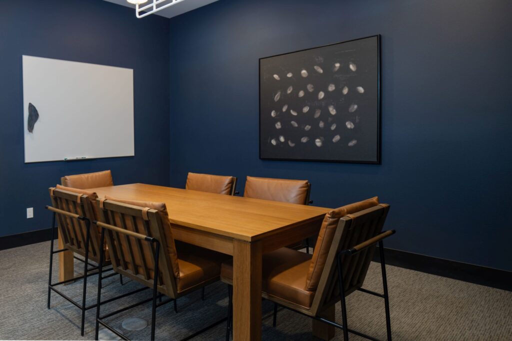 Modern conference room with wood table, leather chairs, whiteboard, and abstract art on a dark blue wall. Office meeting space.